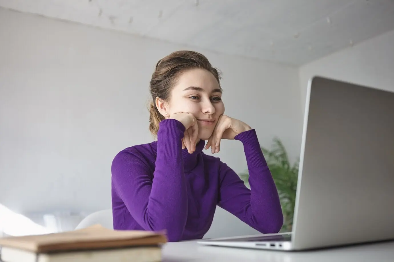 Innenaufnahme einer schönen jungen Frau in einem lilafarbenen langärmeligen Oberteil, die an einem Schreibtisch vor einem geöffneten Laptop sitzt und einen Videoblog ansieht.